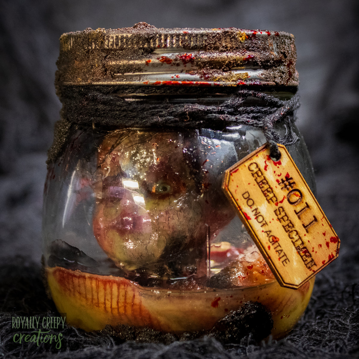 A creepy specimen prop of a severed doll head inside a dirty, rusted glass jar with a dark lid and rope, set against a dark textured background.