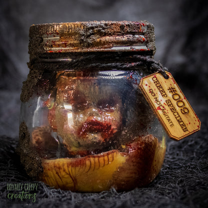 A creepy specimen prop of a severed doll head inside a dirty, rusted glass jar with a dark lid and rope, set against a dark textured background.