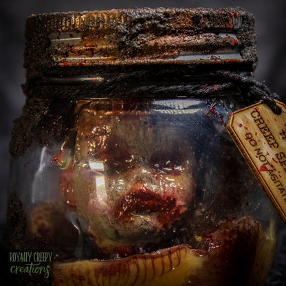 A creepy specimen prop of a severed doll head inside a dirty, rusted glass jar with a dark lid and rope, set against a dark textured background.