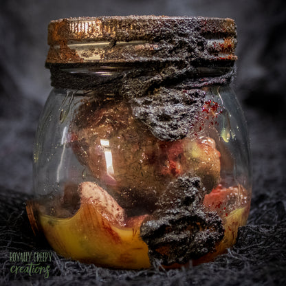 A creepy specimen prop of a severed doll head inside a dirty, rusted glass jar with a dark lid and rope, set against a dark textured background.