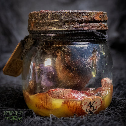A creepy specimen prop of a severed doll head inside a dirty, rusted glass jar with a dark lid and rope, set against a dark textured background.