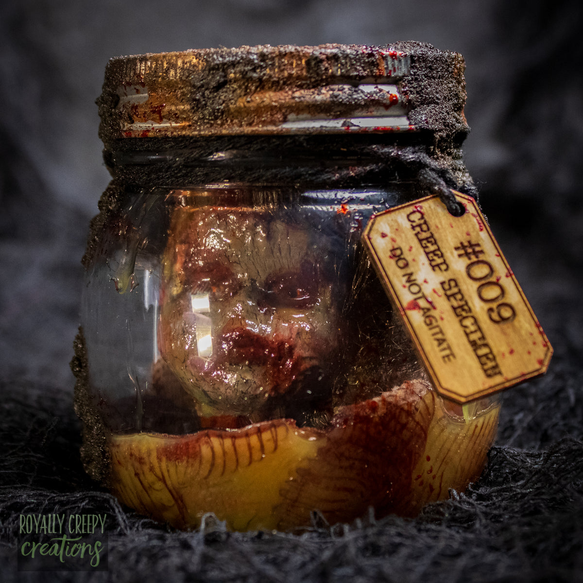 A creepy specimen prop of a severed doll head inside a dirty, rusted glass jar with a dark lid and rope, set against a dark textured background.
