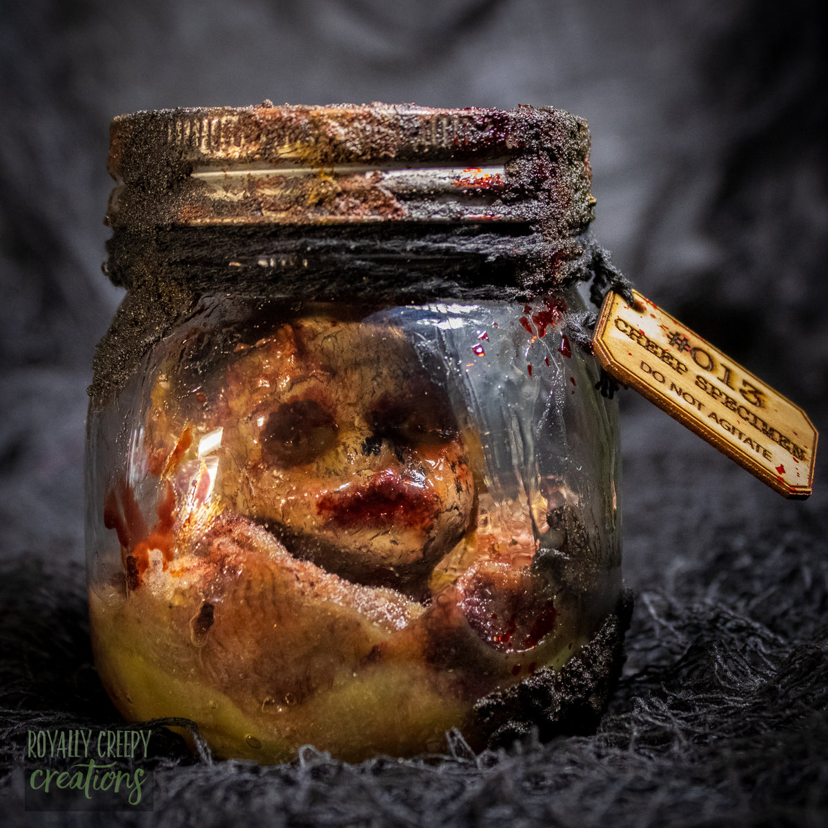A creepy specimen prop of a severed doll head inside a dirty, rusted glass jar with a dark lid and rope, set against a dark textured background.