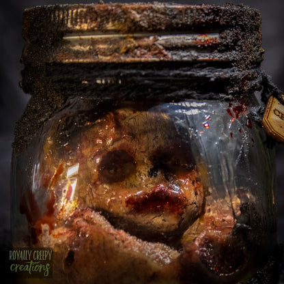 A creepy specimen prop of a severed doll head inside a dirty, rusted glass jar with a dark lid and rope, set against a dark textured background.