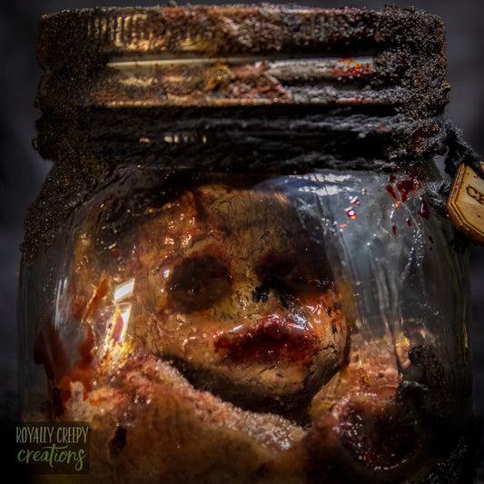 A creepy specimen prop of a severed doll head inside a dirty, rusted glass jar with a dark lid and rope, set against a dark textured background.