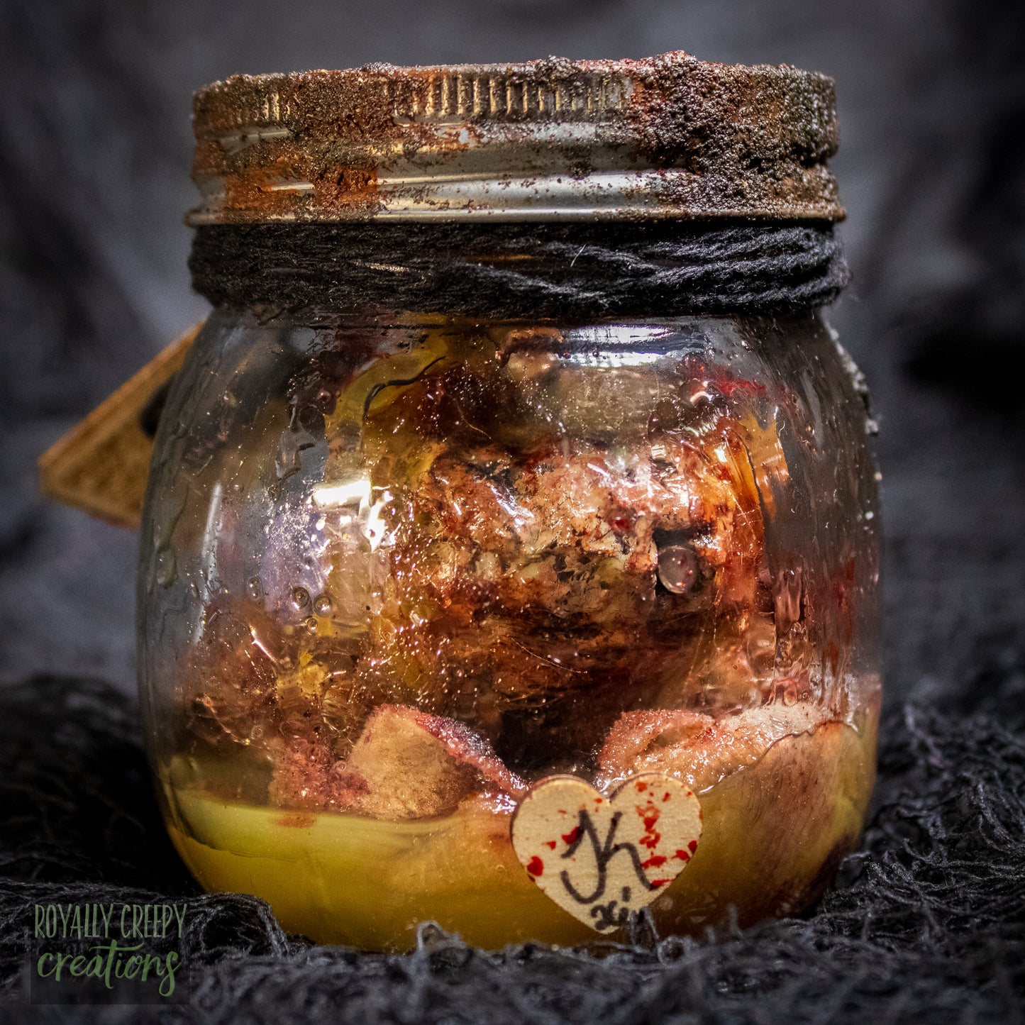 A creepy specimen prop of a severed doll head inside a dirty, rusted glass jar with a dark lid and rope, set against a dark textured background.