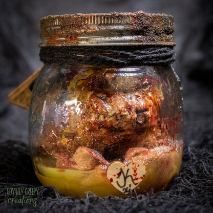 A creepy specimen prop of a severed doll head inside a dirty, rusted glass jar with a dark lid and rope, set against a dark textured background.