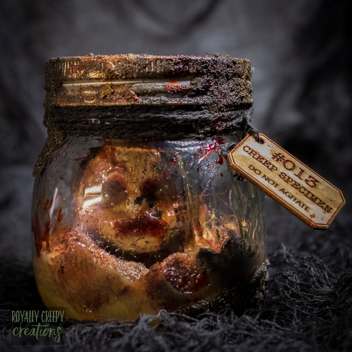 A creepy specimen prop of a severed doll head inside a dirty, rusted glass jar with a dark lid and rope, set against a dark textured background.