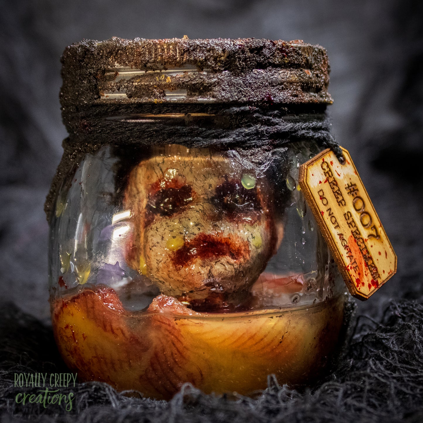 A creepy specimen prop of a severed doll head inside a dirty, rusted glass jar with a dark lid and rope, set against a dark textured background.