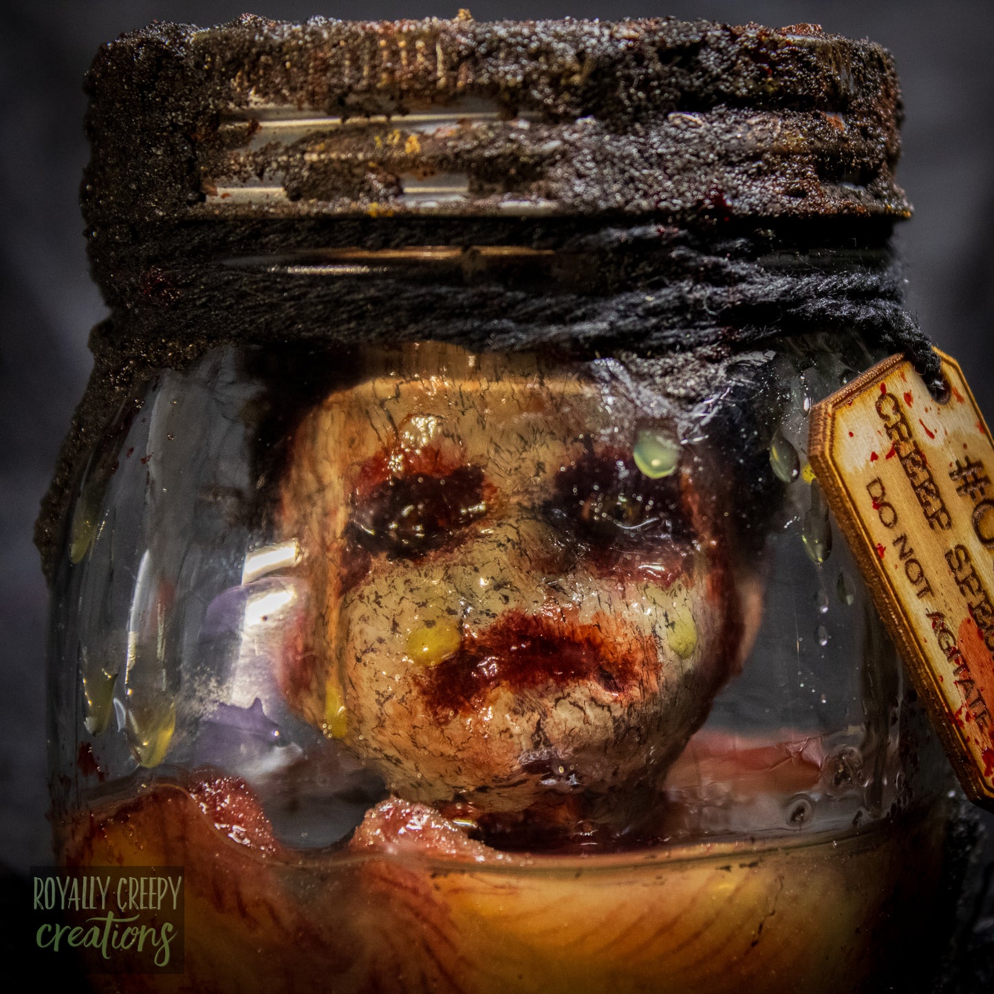 A creepy specimen prop of a severed doll head inside a dirty, rusted glass jar with a dark lid and rope, set against a dark textured background.