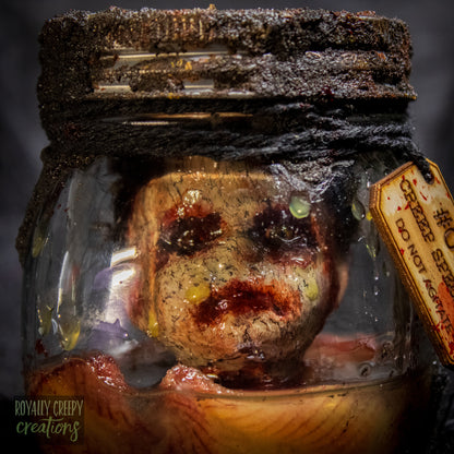 A creepy specimen prop of a severed doll head inside a dirty, rusted glass jar with a dark lid and rope, set against a dark textured background.