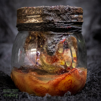 A creepy specimen prop of a severed doll head inside a dirty, rusted glass jar with a dark lid and rope, set against a dark textured background.