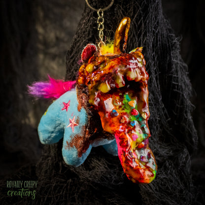 A creepy, horror-themed keychain featuring a plush unicorn vomiting up a rainbow.