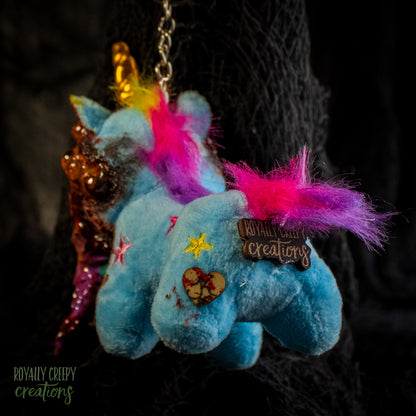 Back view of a blue plush unicorn keychain with a bright pink and purple mane, decorated with a brand tag reading 'Royally Creepy Creations'.