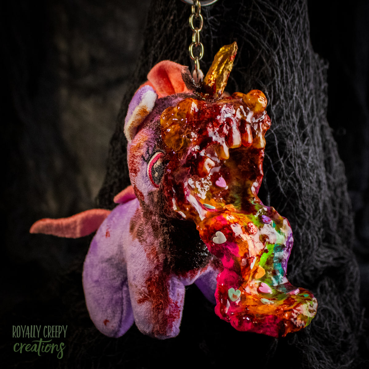 A creepy, horror-themed keychain featuring a plush unicorn vomiting up a rainbow.