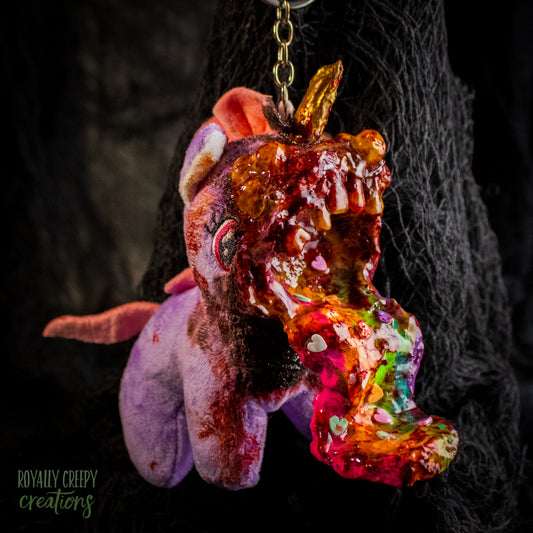 A creepy, horror-themed keychain featuring a plush unicorn vomiting up a rainbow.