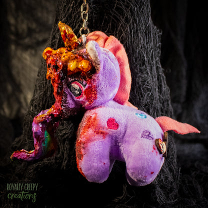 A creepy, horror-themed keychain featuring a plush unicorn vomiting up a rainbow.
