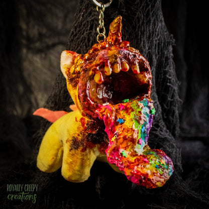 A creepy, horror-themed keychain featuring a plush unicorn vomiting up a rainbow.
