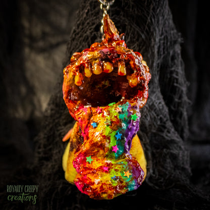 A creepy, horror-themed keychain featuring a plush unicorn vomiting up a rainbow.