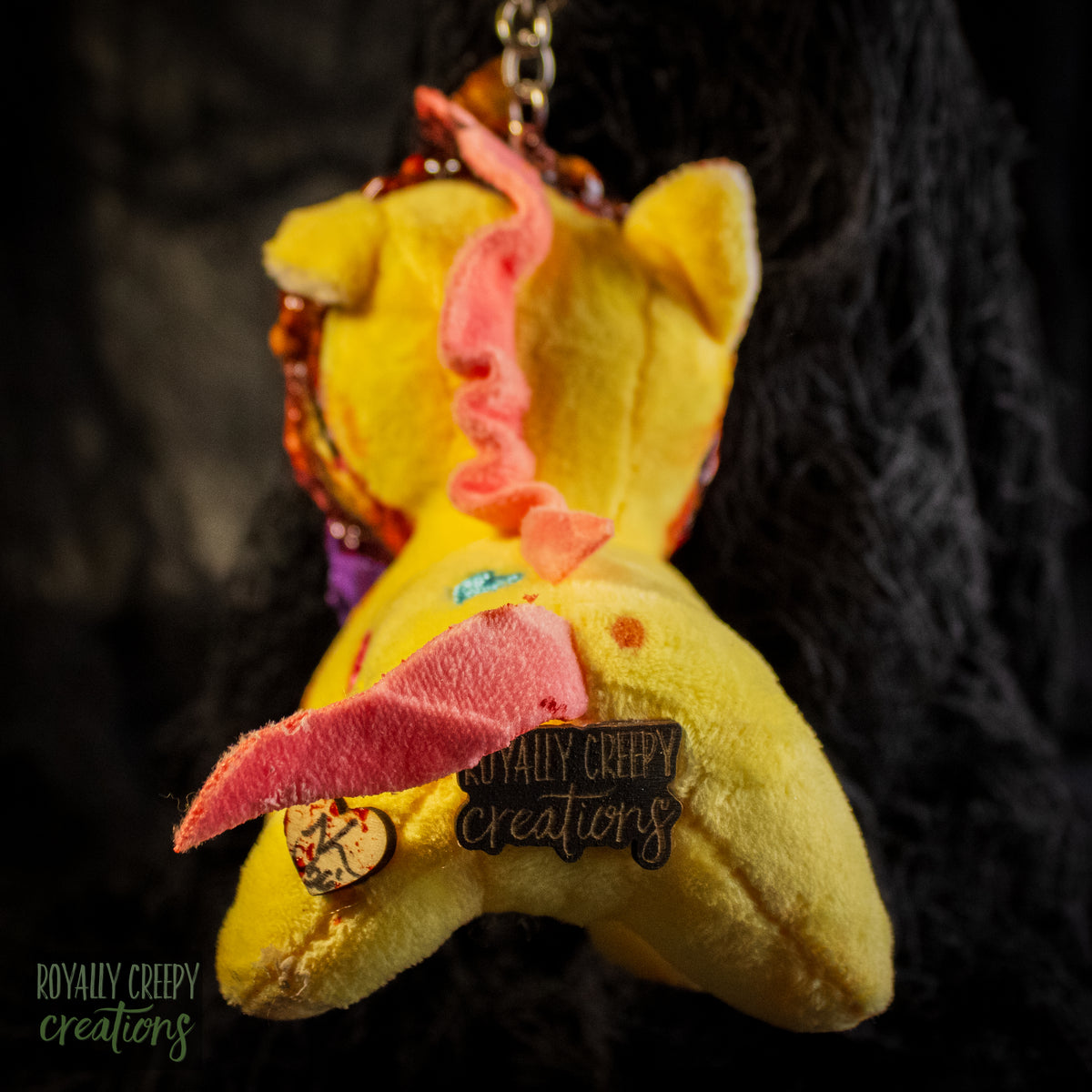 Back view of a yellow plush unicorn keychain with a logo pin for 'Royally Creepy Creations'