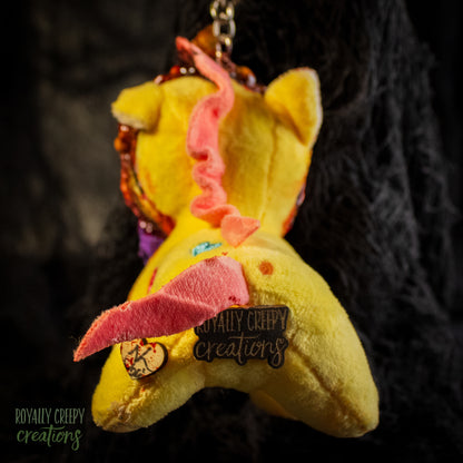 Back view of a yellow plush unicorn keychain with a logo pin for 'Royally Creepy Creations'