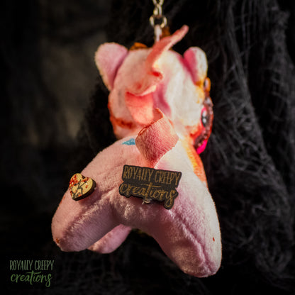 Back view of a pink plush unicorn keychain with a brand pin reading 'ROYALY CREEPY creations'.