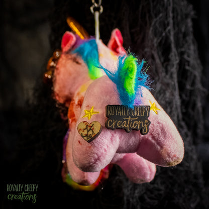 Back view of a pink plush unicorn keychain  with a 'Royally Creepy Creations' and a small heart charm, hanging against a dark textured background.