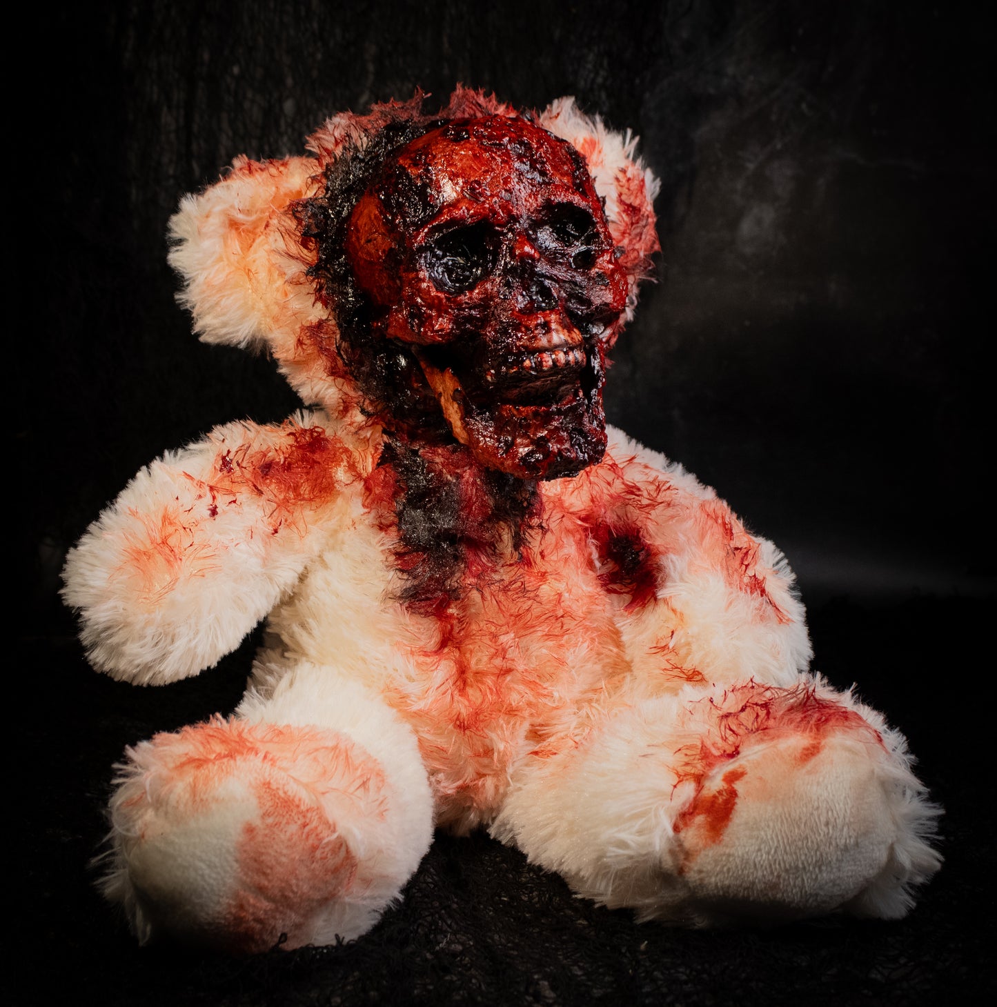 A horror-themed plush teddy bear with an exposed bloody skull for a face, covered in fake blood splatters, set against a dark background.