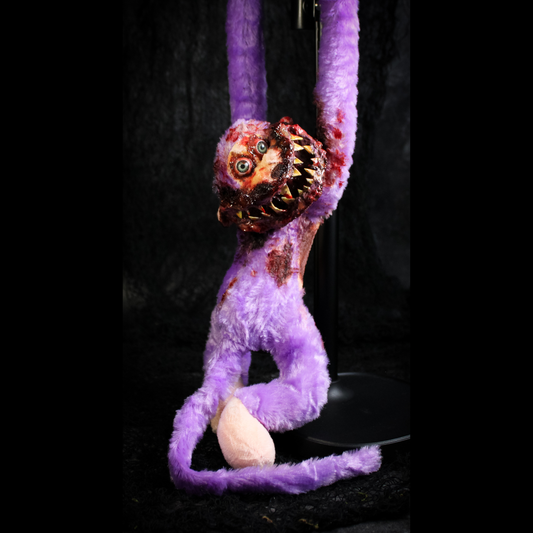 A purple monkey doll with a bloody, toothy mouth and long limbs, posed hanging against a dark studio background.
