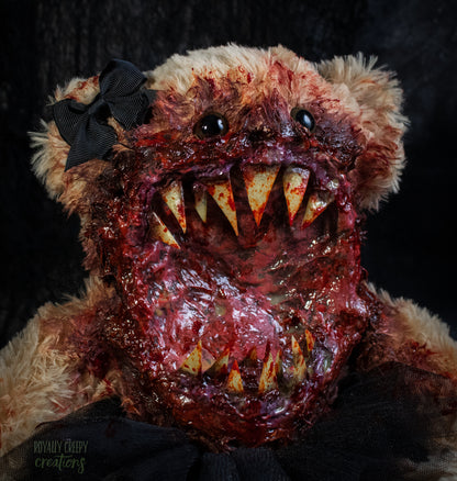 A close-up of a horror-themed plush teddy bear with a bloody, grotesque mouth showing sharp teeth, a black tutu, and a black bow on its head, set against a dark background.