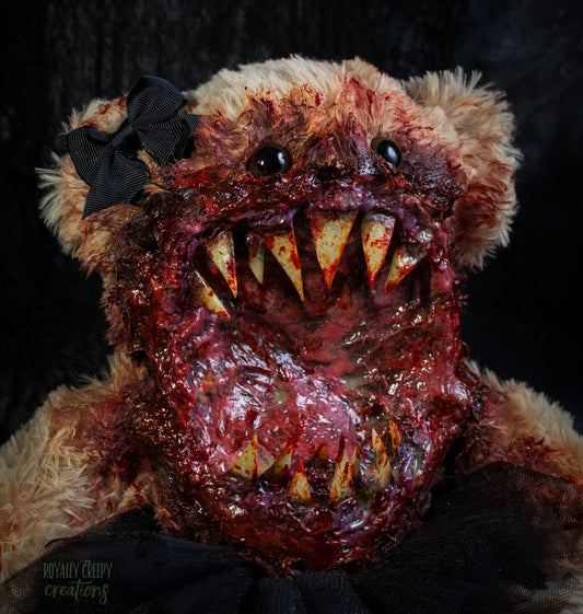 A close-up of a horror-themed plush teddy bear with a bloody, grotesque mouth showing sharp teeth, a black tutu, and a black bow on its head, set against a dark background.