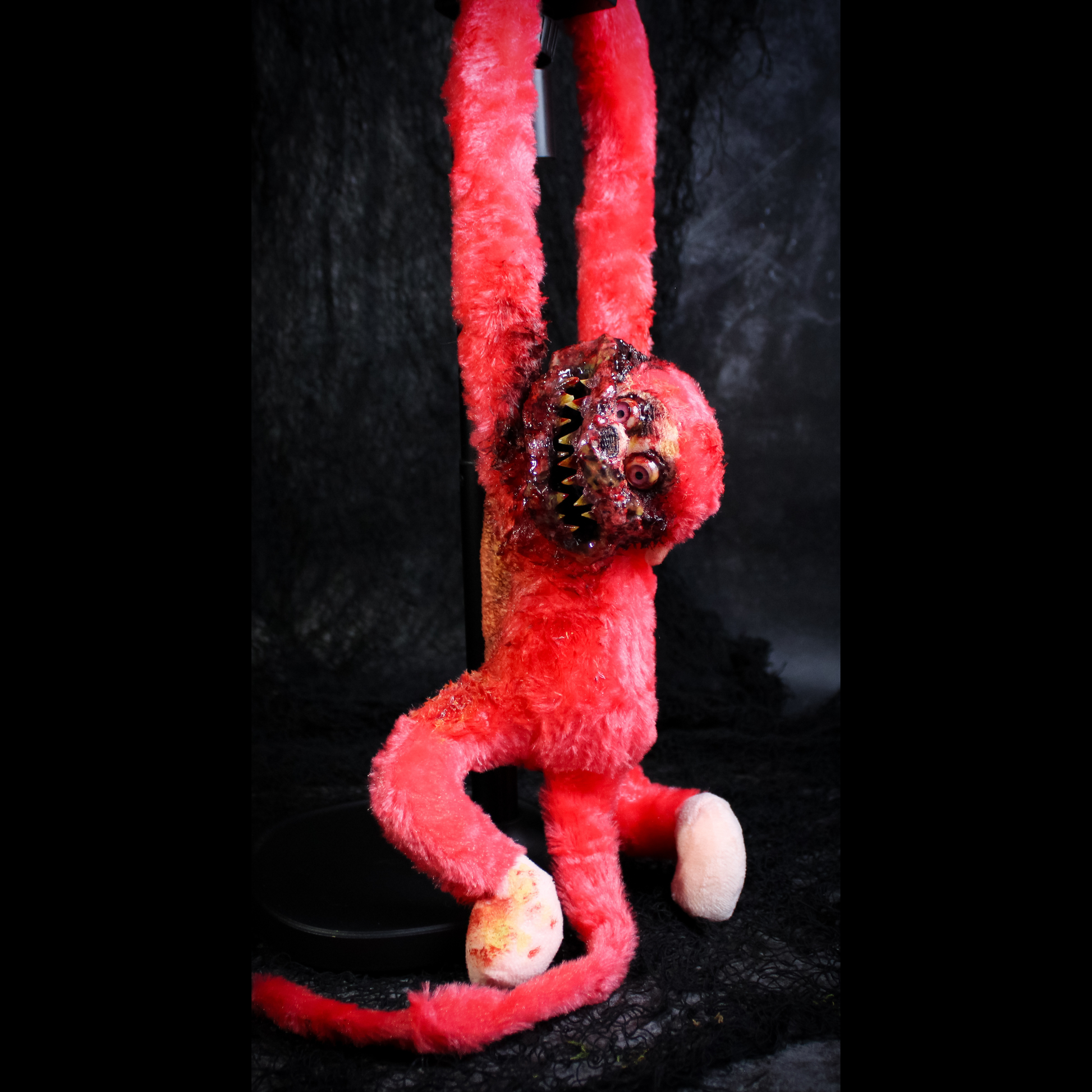 A red plush monkey hanging against a dark textured backdrop. The plush has a horror-style face with large eyes, a wide grin showing teeth.