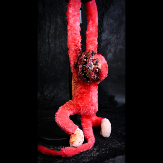 A red plush monkey hanging against a dark textured backdrop. The plush has a horror-style face with large eyes, a wide grin showing teeth.